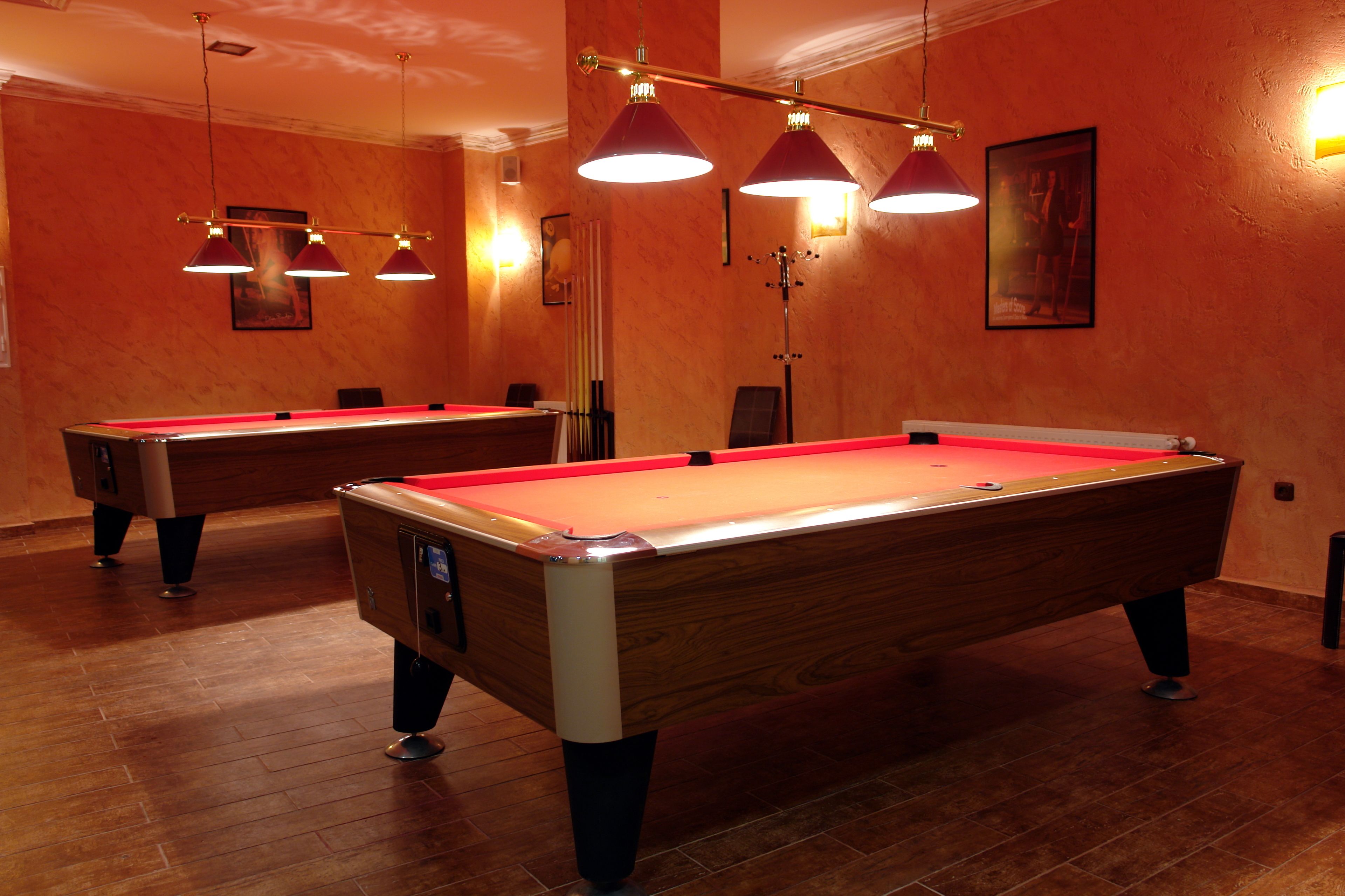 Una mesa de billar con superficie de fieltro rojo, dos mesas de billar e iluminación colgante en una sala con suelo de madera y paredes anaranjadas.