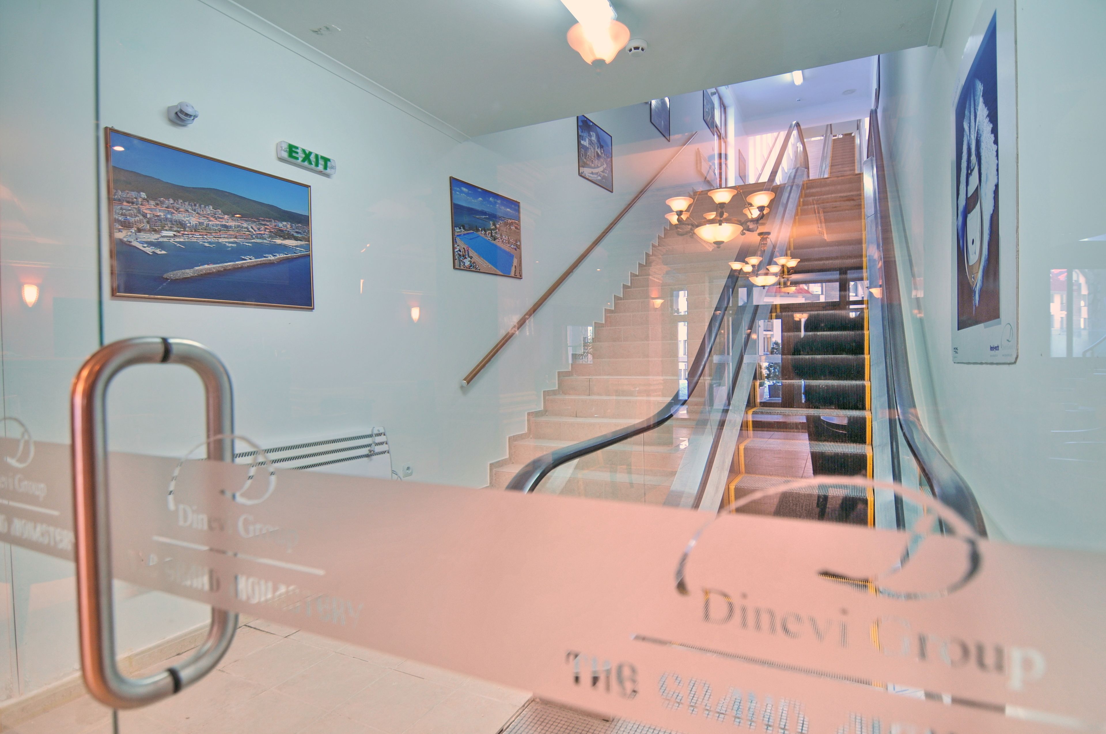 A staircase with an exit sign, glass railings, and framed pictures on the walls.