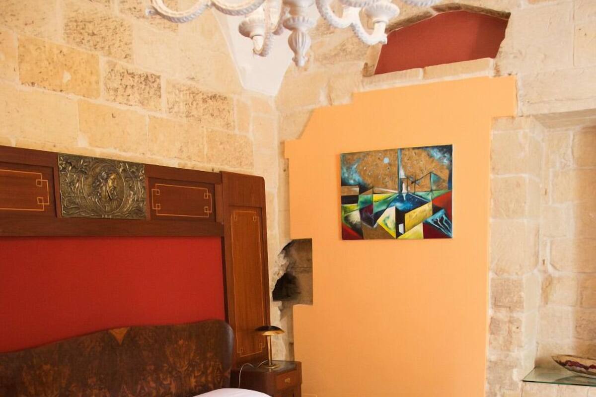 Ein Schlafzimmer mit einem hölzernen Bett, einem roten Kopfteil, einer Chandelüster, einer Steinwand und einem Gemälde.