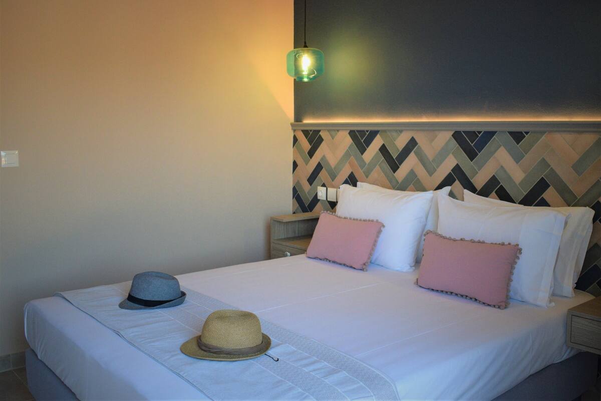 Un lit bien fait, avec du linge de lit blanc et des coussins roses, un chapeau posé sur le lit, et un mur orné d’un motif en chevrons.