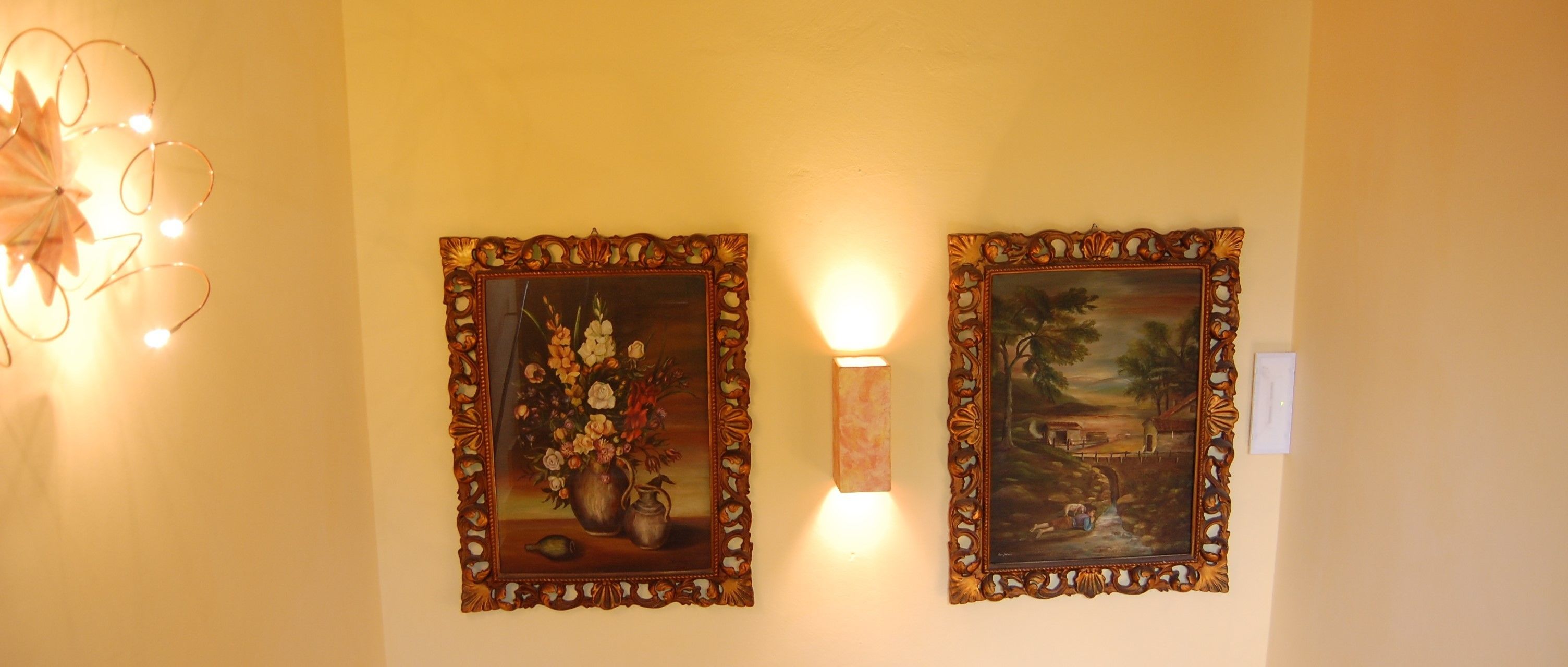 Two ornate framed paintings on a wall, one depicting a floral arrangement and the other a landscape scene.