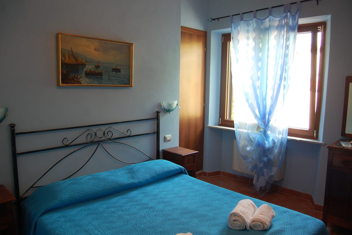 A bedroom with a bed, a painting on the wall, a window with curtains, and a bedside table.