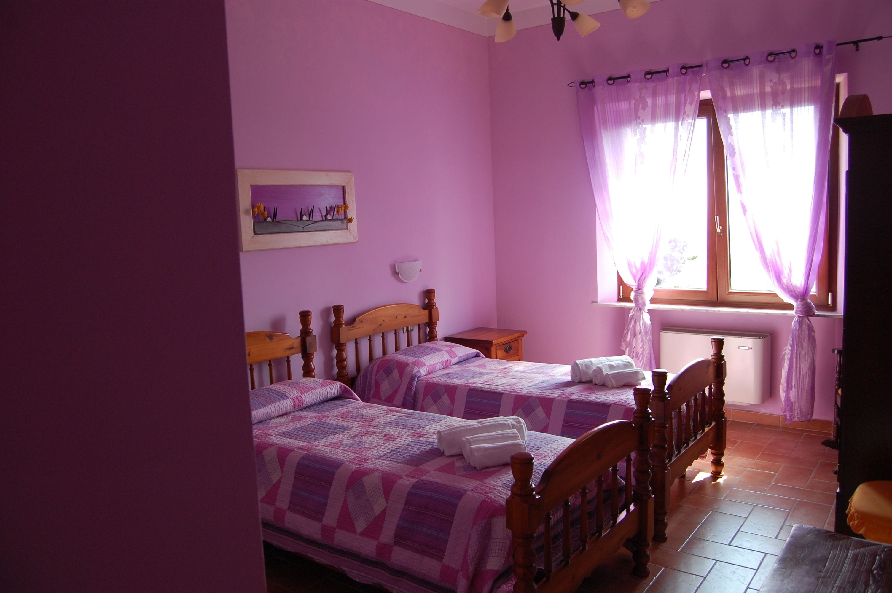 A room with two wooden beds, a small table, a window with purple curtains, and a radiator.