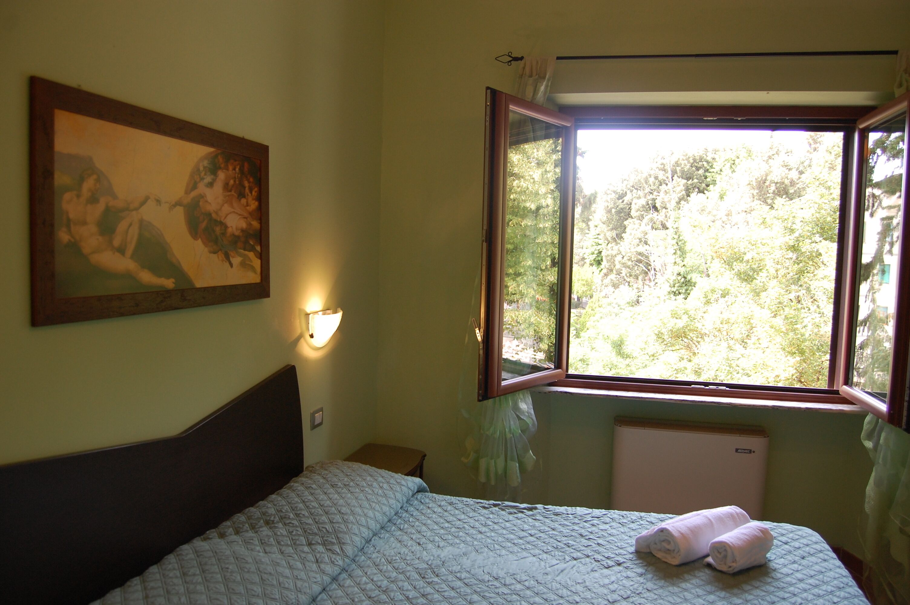 A bedroom with a bed, a painting on the wall, a window with a view of trees, and a wall-mounted light fixture.