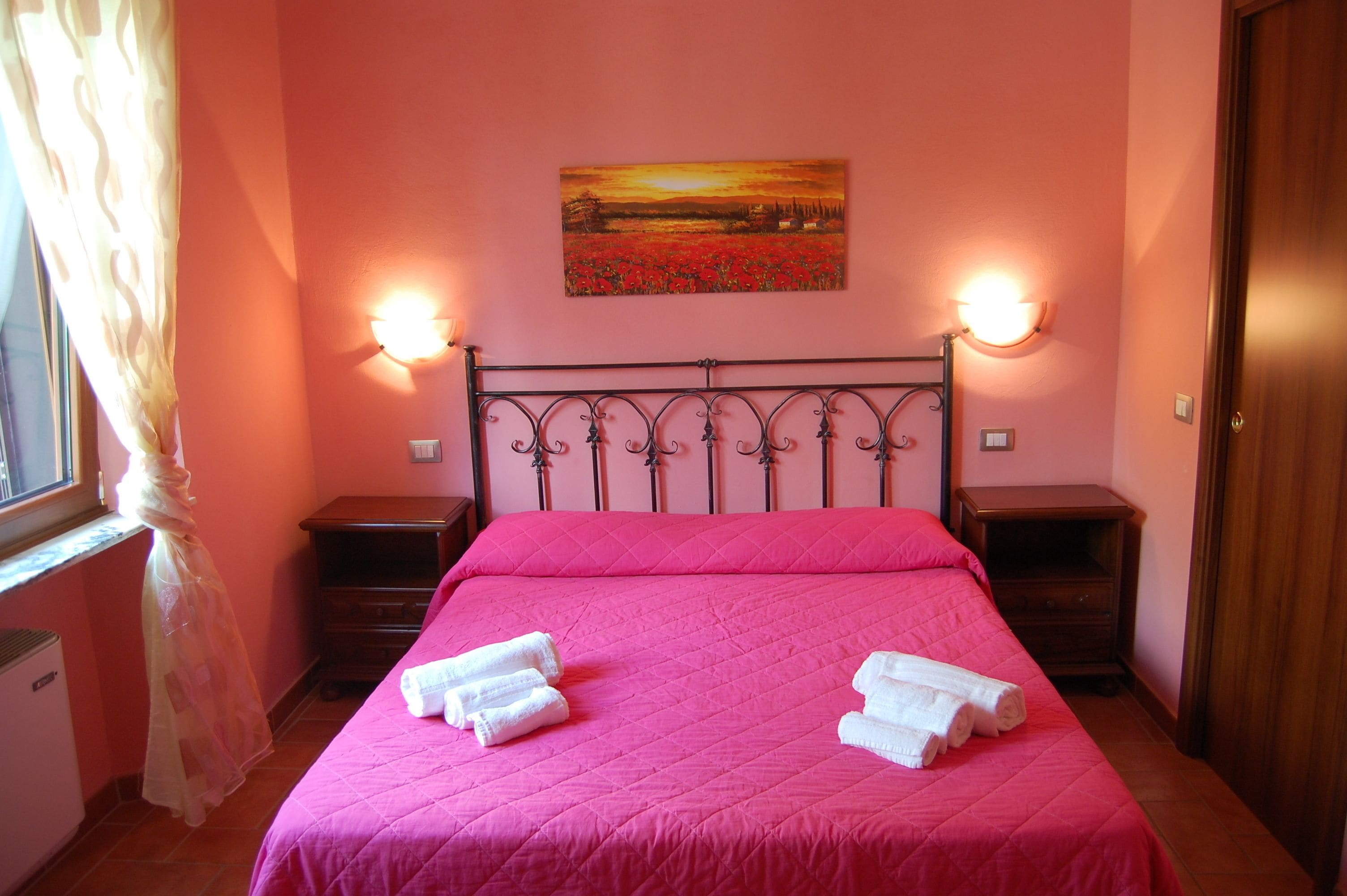 A bedroom with a pink bedspread, two white towels, a painting of a sunset, and two bedside lamps.