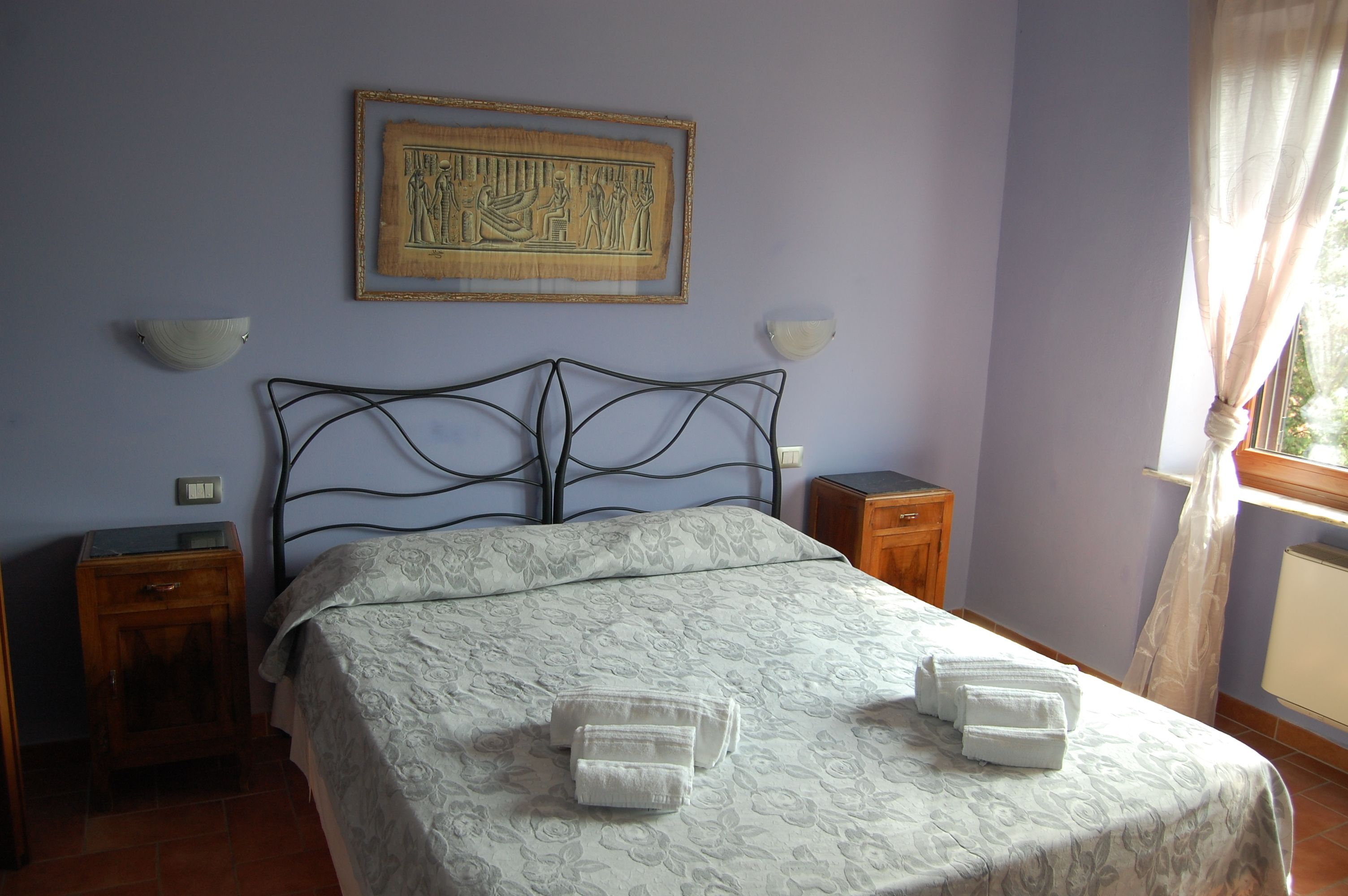 A neatly made bed with a patterned bedspread and two towels on the side. There is a wall-mounted picture above the bed and two bedside tables with lamps.