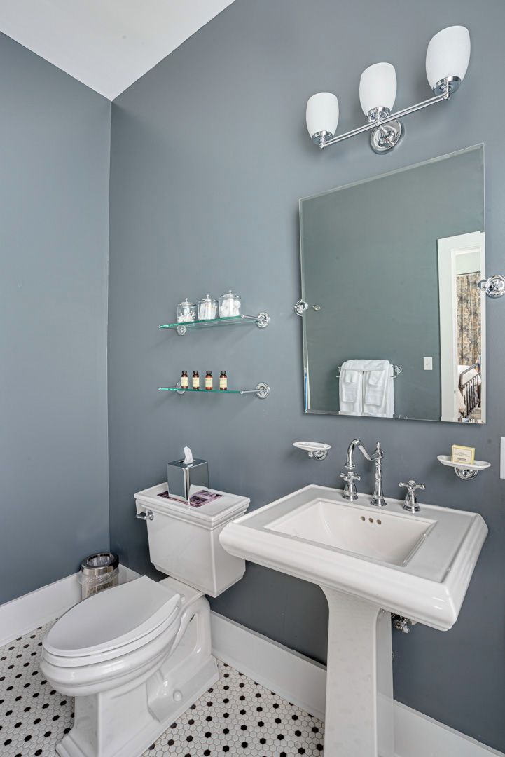 A bathroom with a toilet, sink, and mirror. There are shelves with toiletries and a trash can.