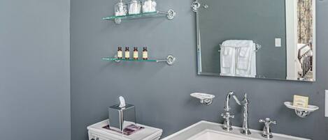 A bathroom with a toilet, sink, and mirror. There are shelves with toiletries and a trash can.