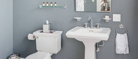 A bathroom with a toilet, sink, and mirror. There are shelves with toiletries and a towel rack.
