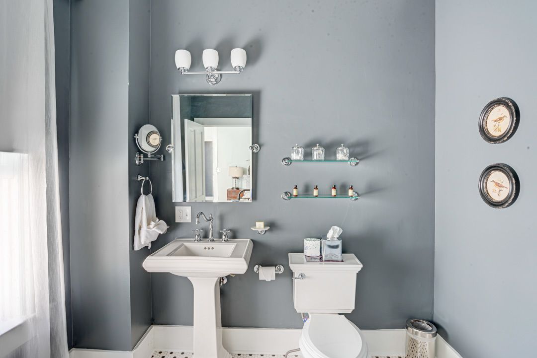 A bathroom with a pedestal sink, a toilet, a mirror, and two shelves with toiletries.