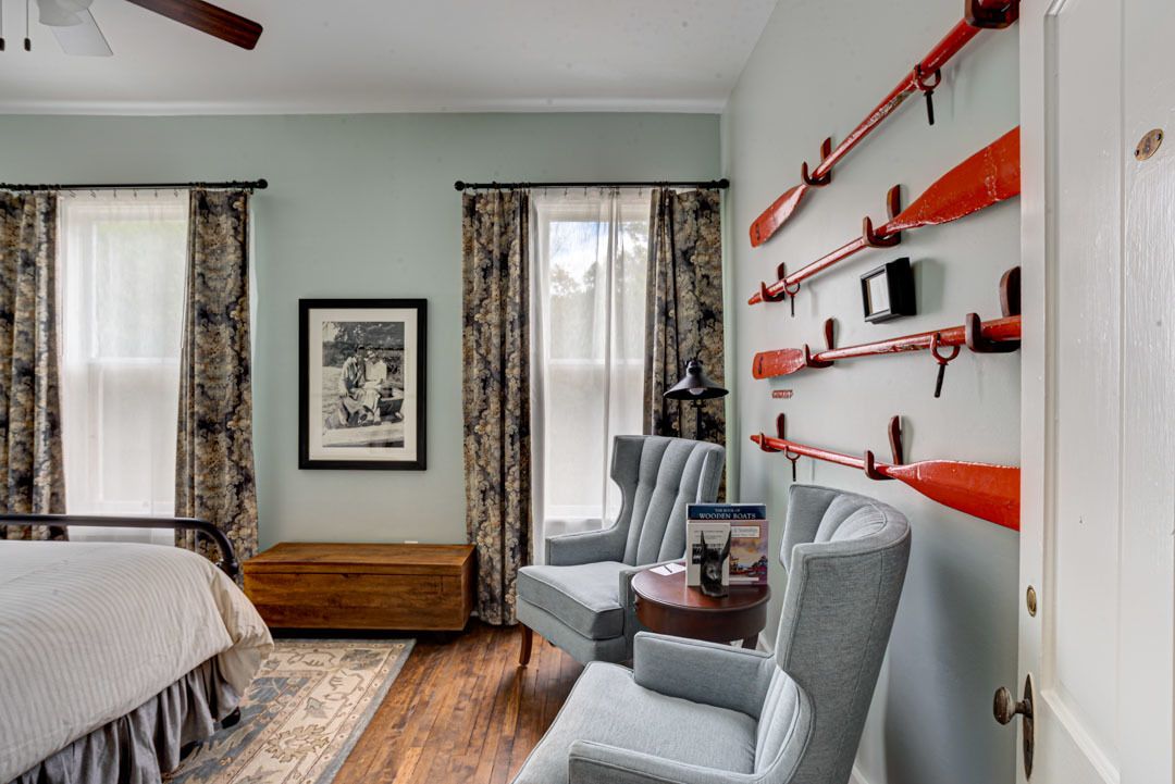 A bedroom with a bed, two armchairs, a small table, a framed picture, and a wall with red objects.