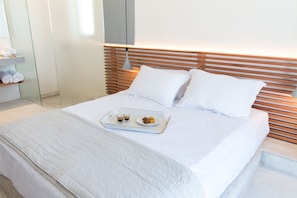 A neatly made bed with a tray of food and drinks, a wooden headboard, and a white wall with a shelf holding towels. - Wabi Luxury Suites (Ios)