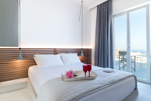 Habitación de hotel con una cama grande, una bandeja con una flor y dos vasos rojos, y vista al exterior.