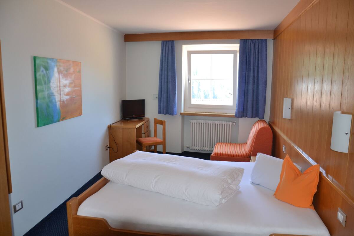 Hotellihuone, jossa on sänky, työpöytä, jossa on tietokone, tuoli ja ikkuna, jossa on verhot.