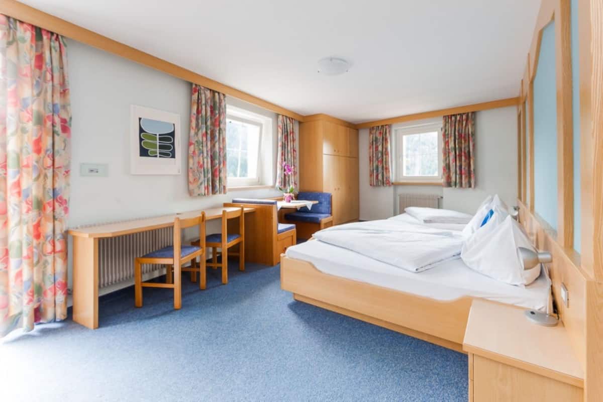 Hotellihuoneessa on sänky, työpöytä ja tuoli. Huoneessa on ikkuna, jossa on verhot, patteri ja seinällä taulu.