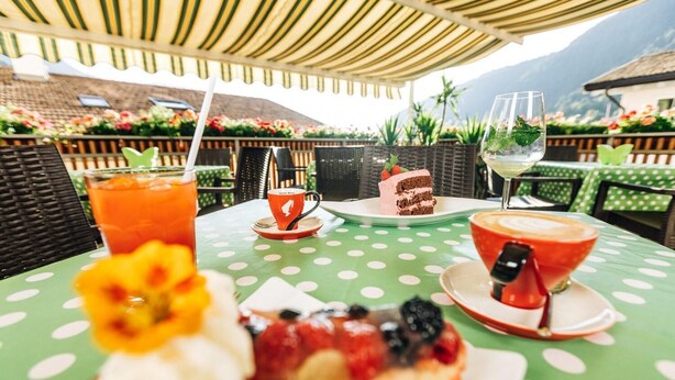 A table with a polka dot tablecloth, a slice of cake, a glass of iced tea, a glass of white wine, and a dessert with berries and whipped cream. - Zum Löwen-Post Hotel (Trodena)