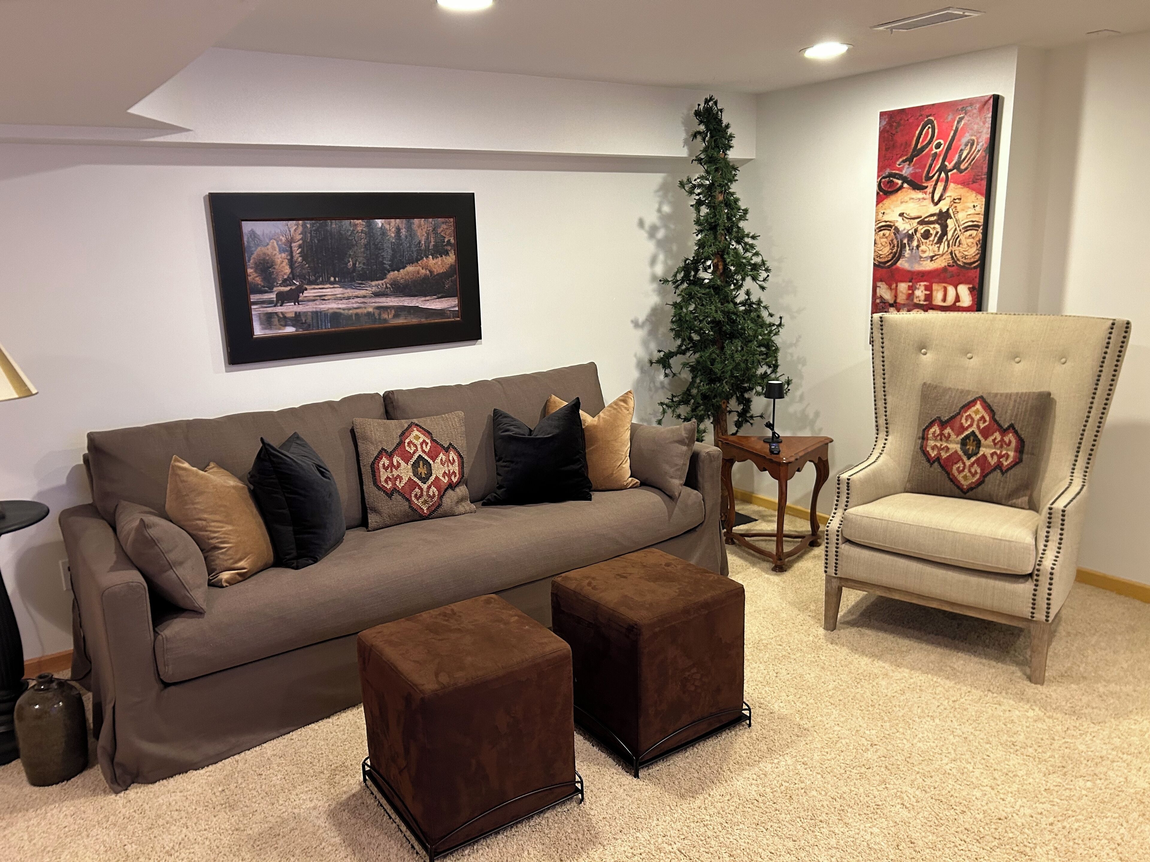 Un salon avec un canapé marron, deux poufs, un grand sapin de Noël, un tableau encadré au mur et un fauteuil avec un coussin.