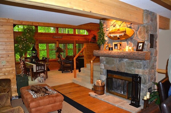 A cozy lodge interior with a stone fireplace, wooden dining area, and a view of the forest through large windows.