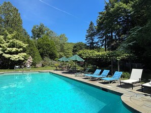 Piscina azul límpida com espreguiçadeiras e guarda-sóis, rodeada por árvores.