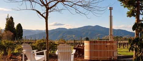 A rustic outdoor seating area with two white chairs and a wooden barrel, set against a backdrop of mountains and vineyards.