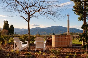 A rustic outdoor seating area with two white chairs and a wooden barrel, set against a backdrop of mountains and vineyards.