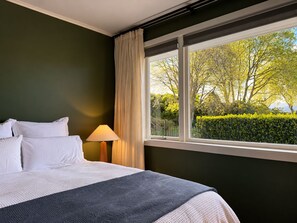 A hotel room with a bed, a lamp, and a window with a view of trees and bushes.