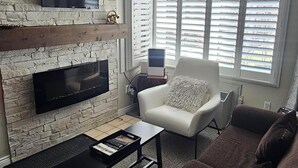 A living room with a stone wall, a fireplace, a flat-screen TV, a white armchair, a brown sofa, and a window with blinds.