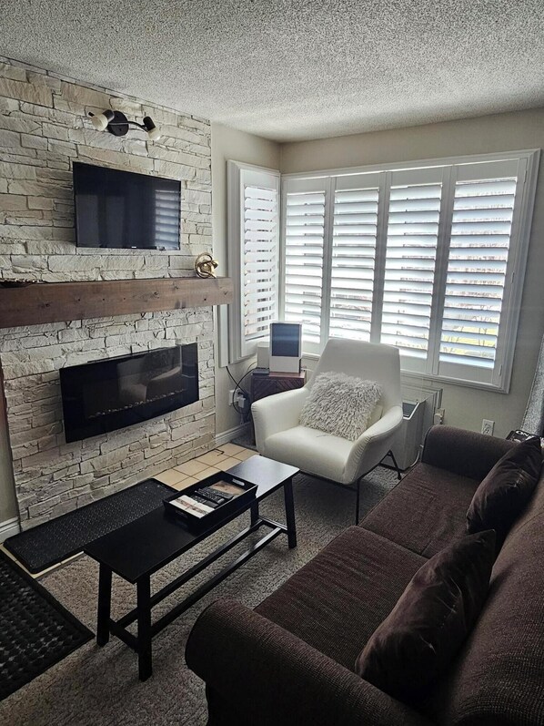 A living room with a stone wall, a fireplace, a flat-screen TV, a white armchair, a brown sofa, and a window with blinds.