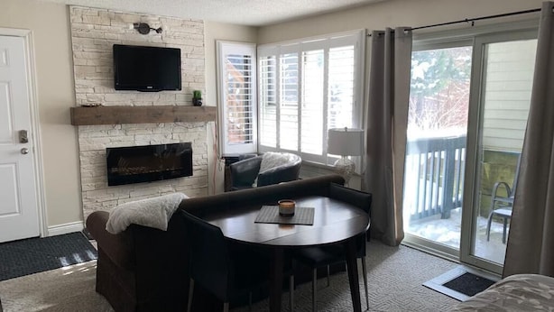 A cozy living room with a fireplace, a flat-screen TV mounted on a stone wall, a dining table with chairs, and a sliding glass door leading to a balcony.