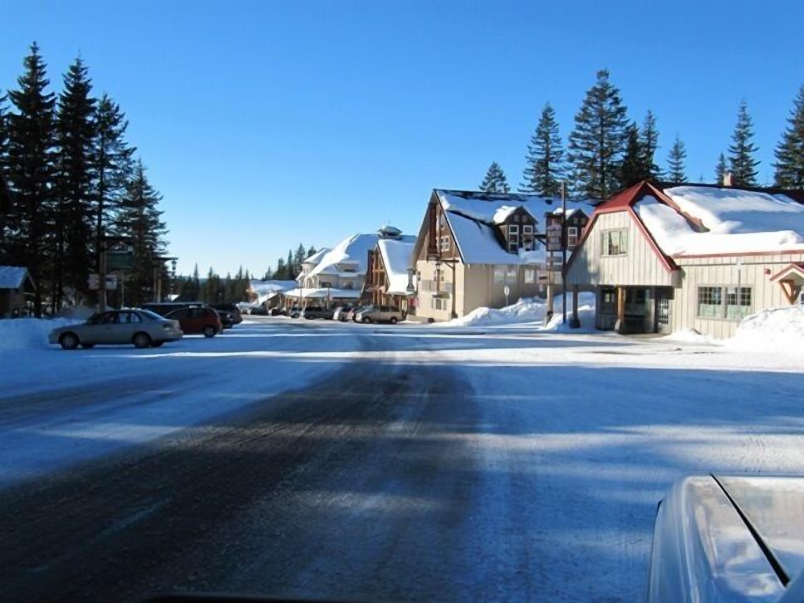 A snow-covered street with traditional wooden buildings, a clear sky, and parked cars.