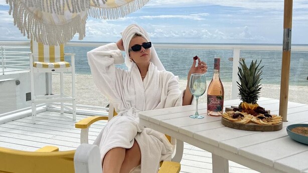 Eine Person sitzt an einem weißen Tisch mit einem gestreiften Strandsonnenschirm, einer Getränkeflasche und einer Obstplatte.