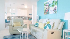 Una sala de estar con un sofá, sillones y una mesita. Se ve una cocina al fondo, con un revoque de azulejos.