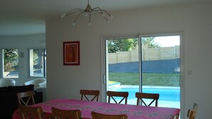 Un coin repas avec une table recouverte d’une nappe à motifs floraux, entourée de chaises en bois, et une vue sur une piscine à travers une porte-fenêtre coulissante.