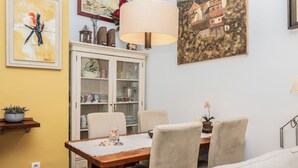 A dining area with a wooden table, beige chairs, a white cabinet with glass doors, and a painting of a house on the wall. - Lovely apartment in the historic district of Seville (+ WiFi & AC) (Sevilla)