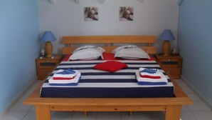 A neatly made bed with striped bedding, two pillows, and folded towels on a wooden bed frame. There are two bedside tables with lamps and a small decorative item on each. The room has a tiled floor and a simple, clean design. - A Deshaies F5 sea view equipped for families / groups nearby beaches   (DESHAIES)