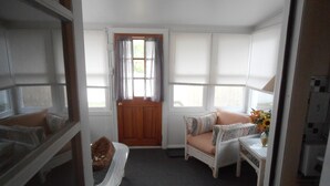 A room with a wooden door, a cushioned bench, a wicker chair, a small table with a vase, and a window with blinds.