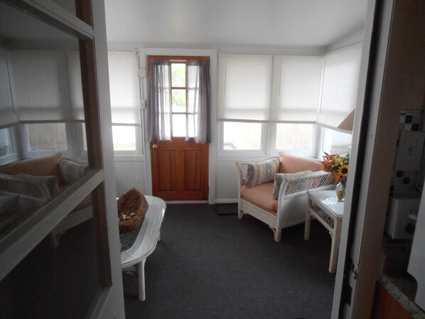 Una habitación con puerta de madera, un banco acolchado, una silla de mimbre, una mesita con un jarrón y una ventana con persianas.