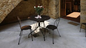 A stone-walled courtyard with a round table and four chairs, a vase with flowers, and a wooden bench.