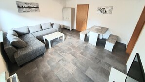 A modern living room with a grey sofa, a white coffee table, and a small white table with benches. There is a wooden door and a white cabinet in the background.