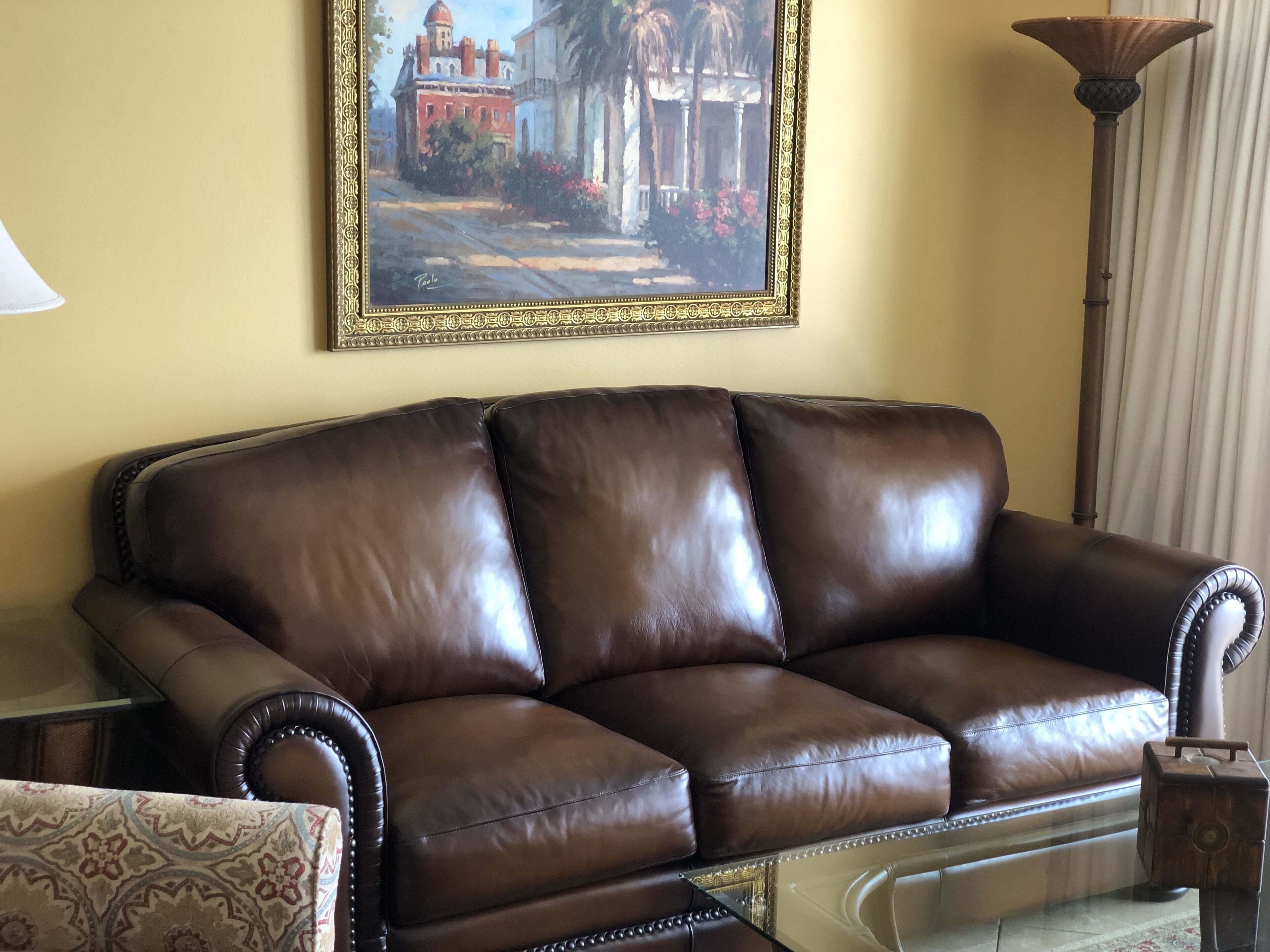 A brown leather sofa with a glass coffee table in front, a patterned armchair, and a painting of a building on the wall.