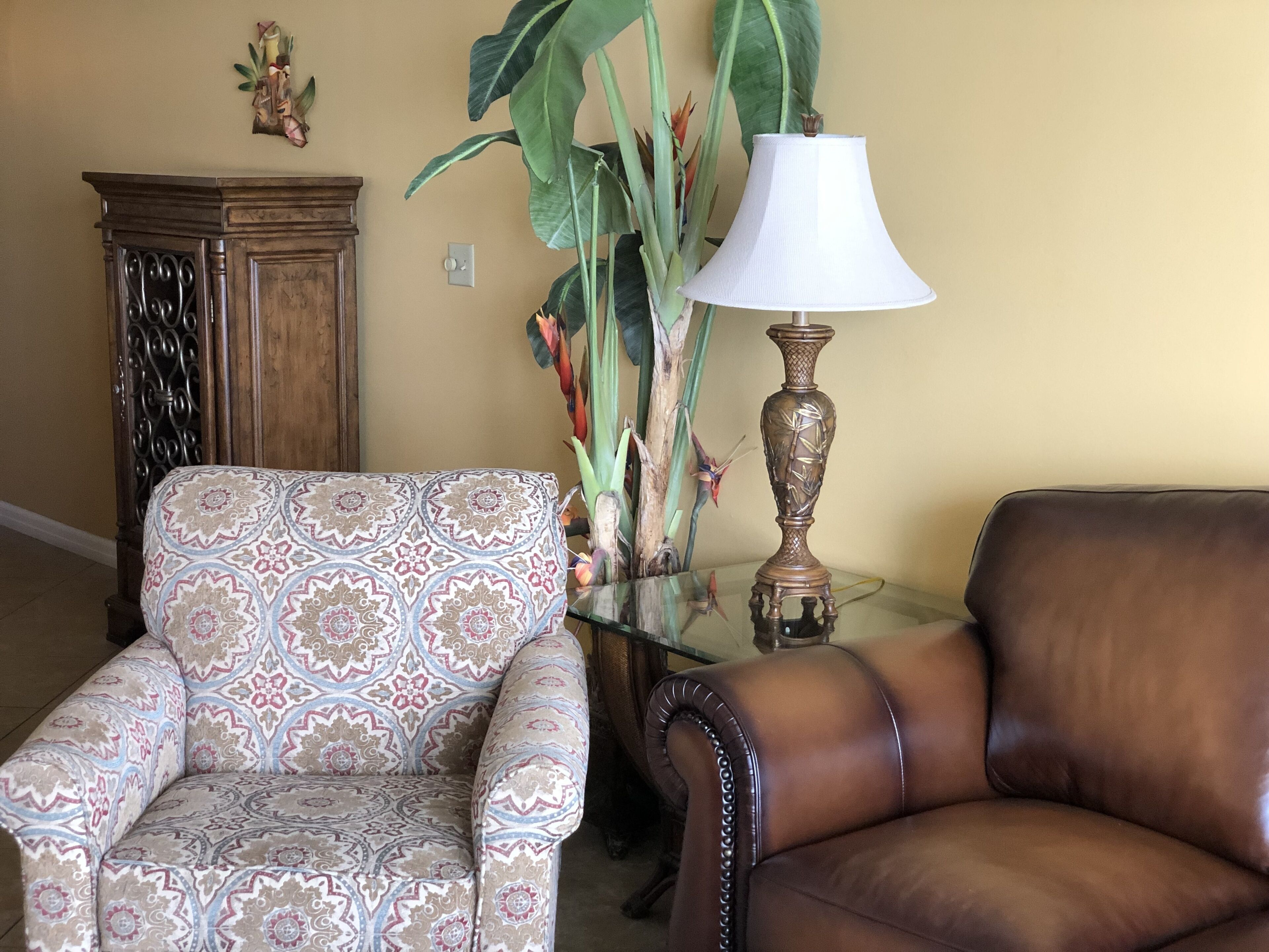 Uma sala de estar com uma poltrona estampada, um sofá de couro, uma mesa de centro de vidro, um abajur decorativo e um armário de madeira.