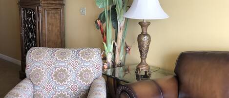 Uma sala de estar com uma poltrona estampada, um sofá de couro, uma mesa de centro de vidro, um abajur decorativo e um armário de madeira.