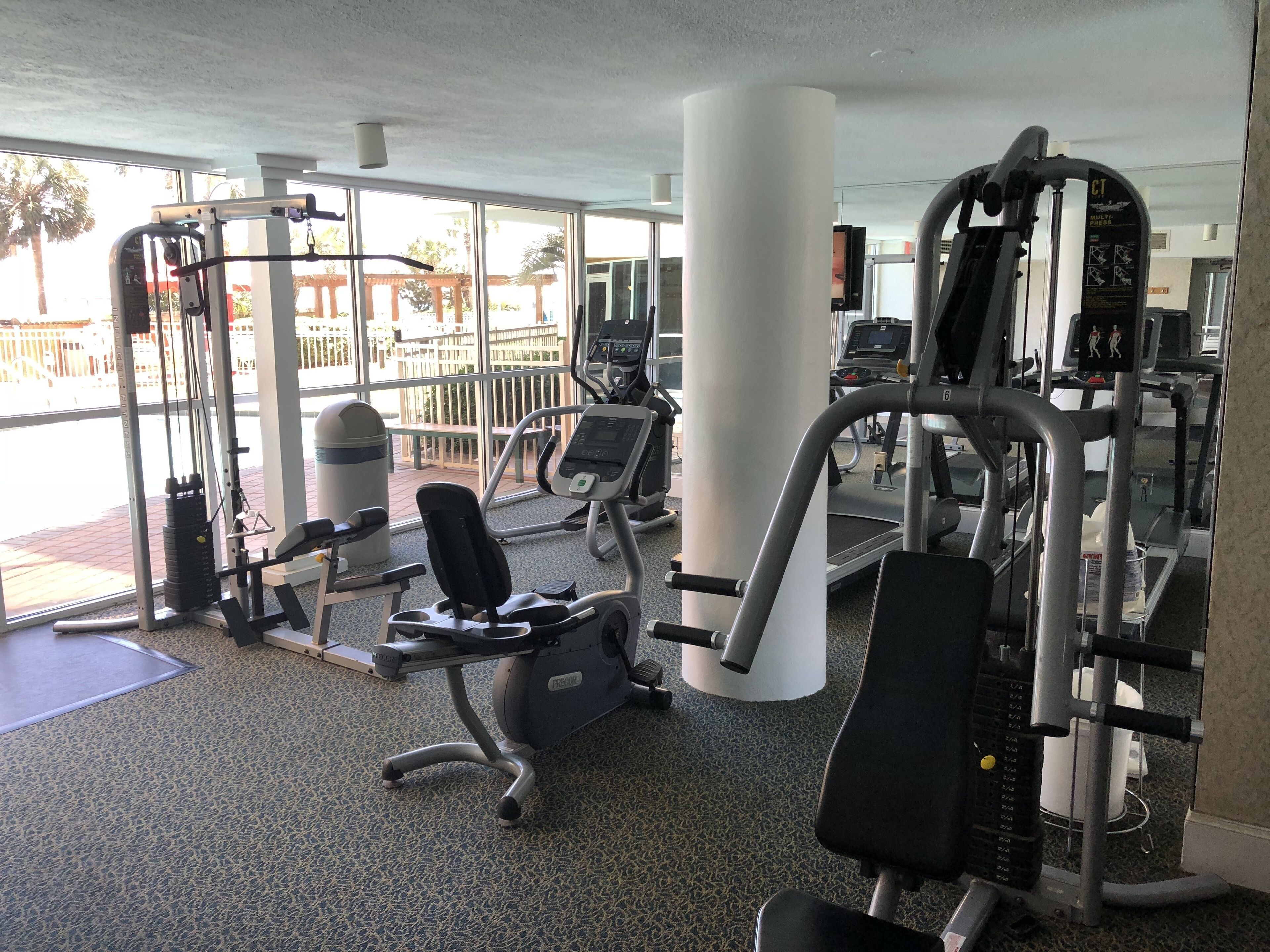 A gym with various exercise machines, large windows, and a view of palm trees outside.