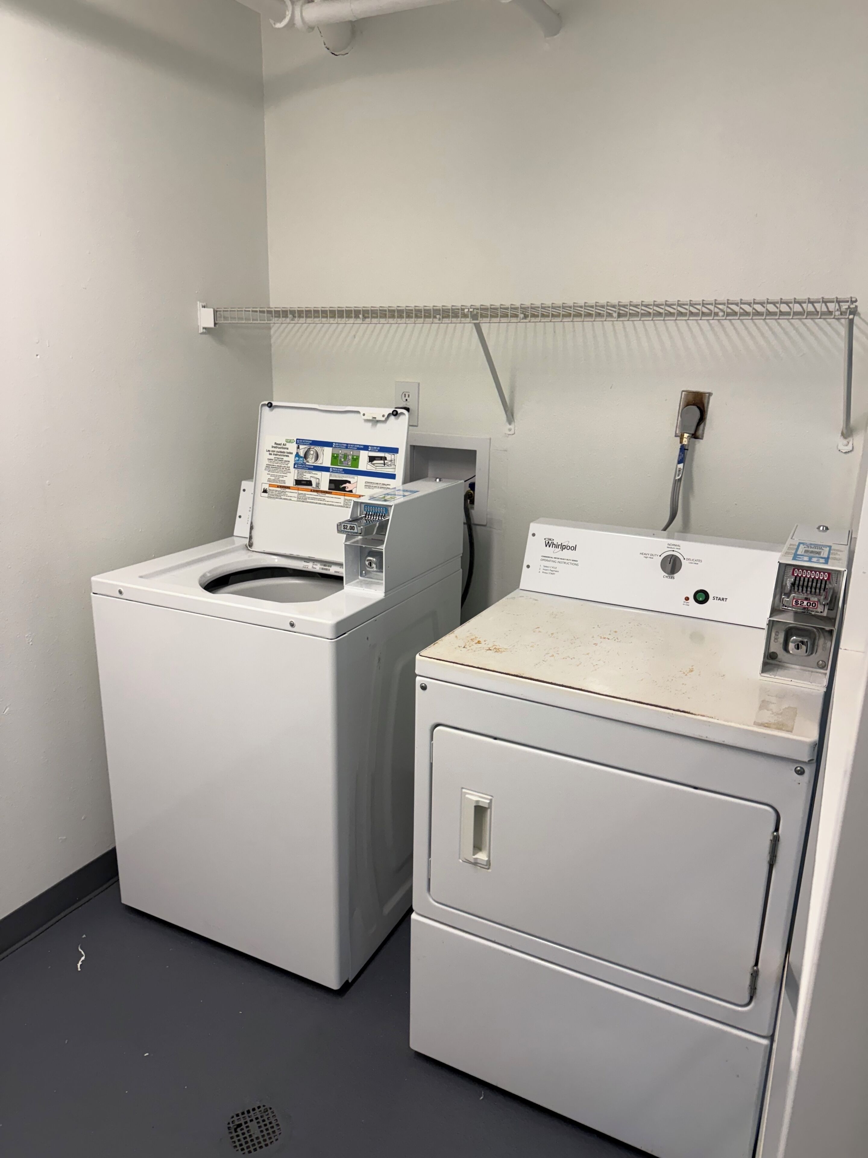 A laundry area with a front-loading washer and dryer, a metal shelf above the dryer, and a power outlet on the wall.