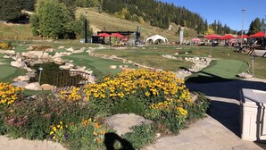 Un campo de golf con un estanque, edificios de techo rojo y montañas al fondo.