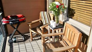 Un conjunto de mesa y silla de madera en un balcón, con una parrilla roja y equipo de limpieza cerca.