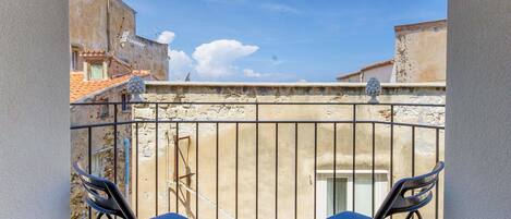 Un balcón con dos sillas azules, piso de baldosas y vista a edificios bajo un cielo despejado.