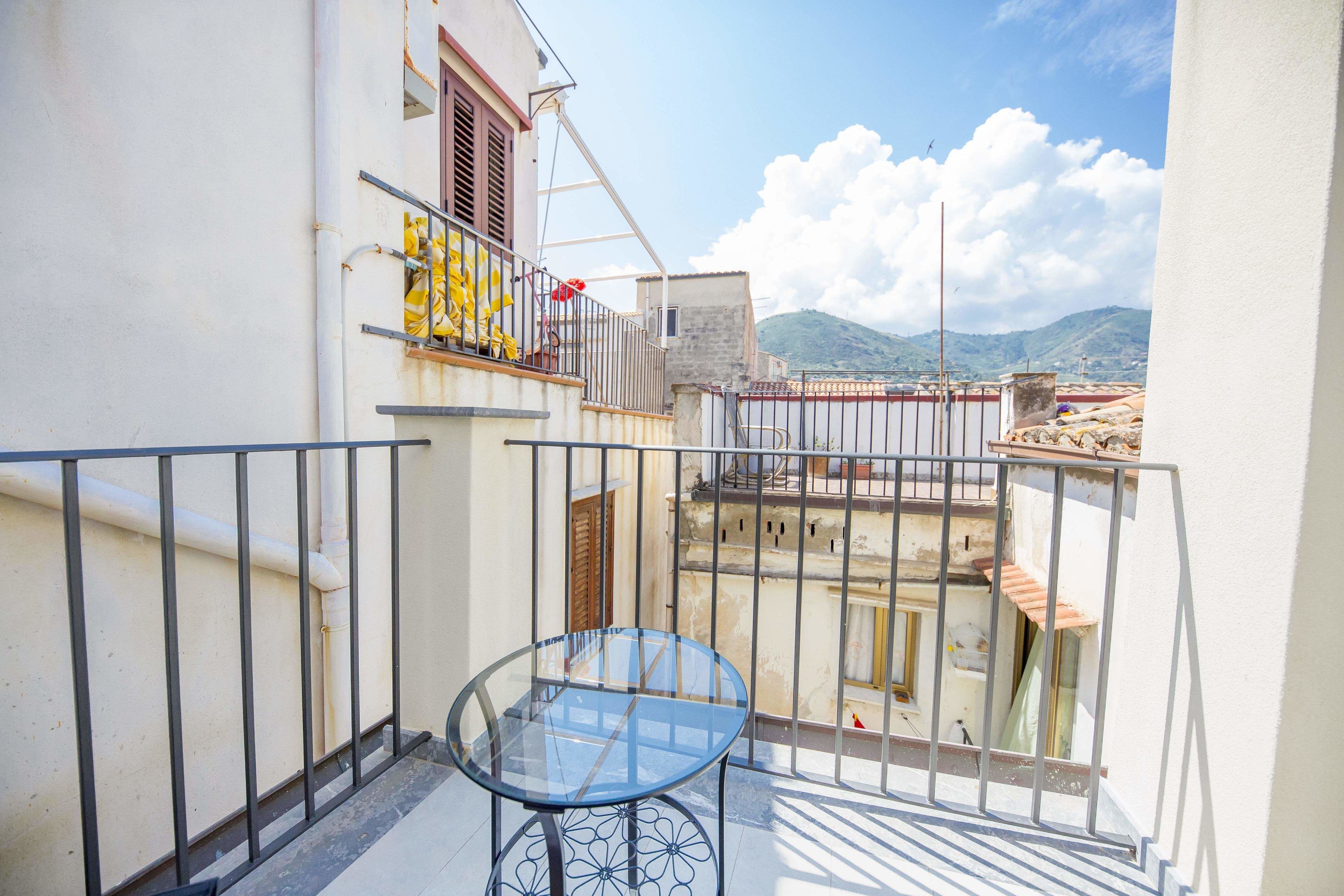 A balcony with a glass table and metal railing, overlooking a cityscape with buildings and mountains in the distance.