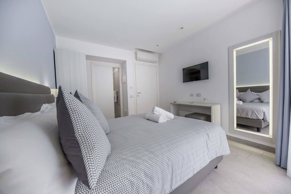 Una habitación de hotel moderna con una cama bien hecha, una mesita de noche, un televisor montado en la pared y un espejo grande con marco iluminado.