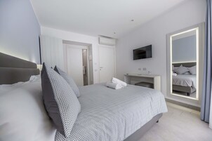 Una habitación de hotel moderna con una cama bien hecha, una mesita de noche, un televisor montado en la pared y un espejo grande con marco iluminado.